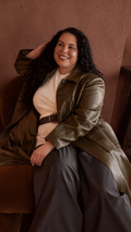 Modal wearing an olive green jacket over a cream top, paired with  charcoal colour pants and a wide black leather belt with metal eyelets, sitting on a brown velvet chair.
