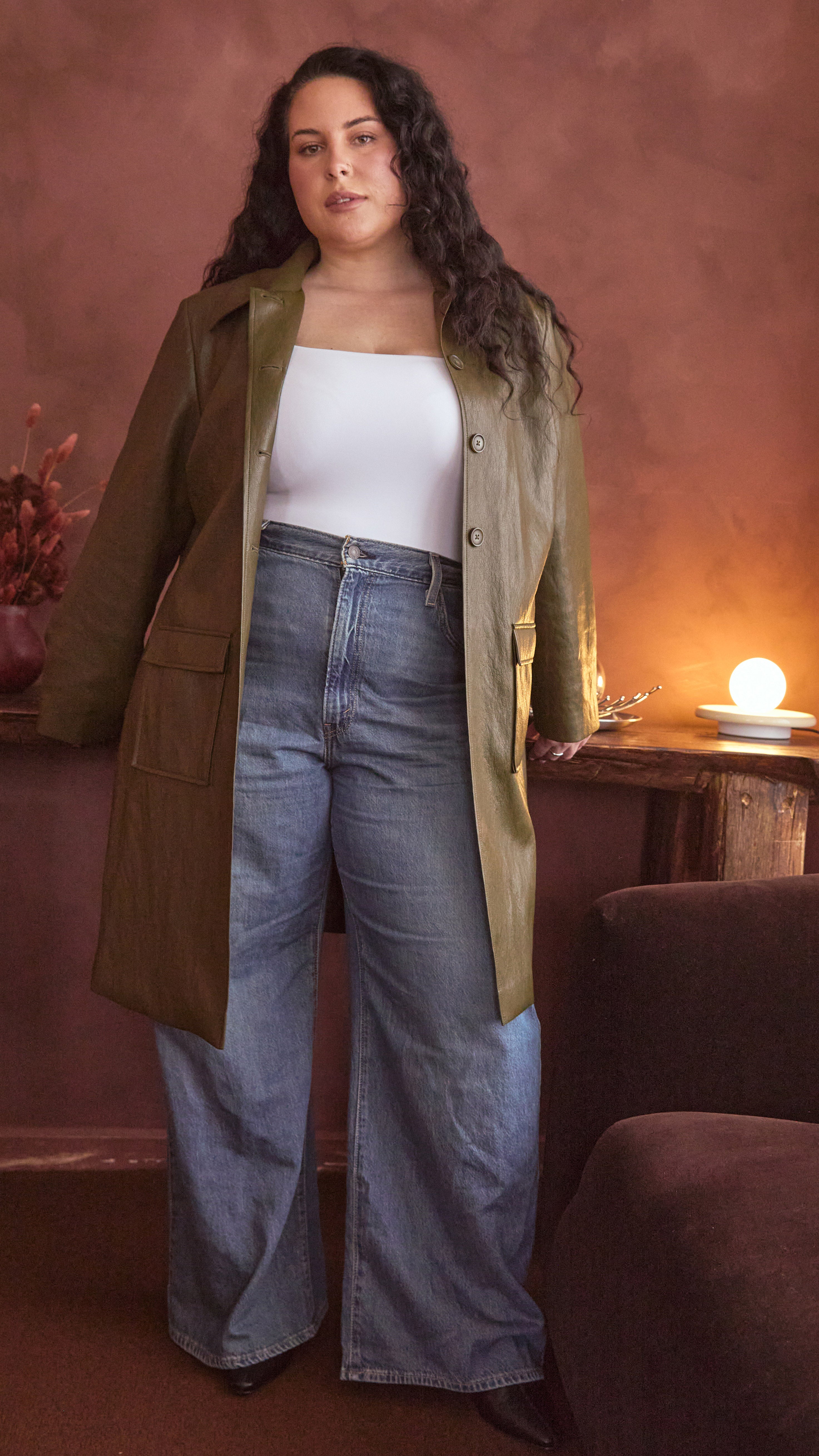 Modal wearing an olive green jacket over a white bodysuit and blue jeans in a warm-toned room.