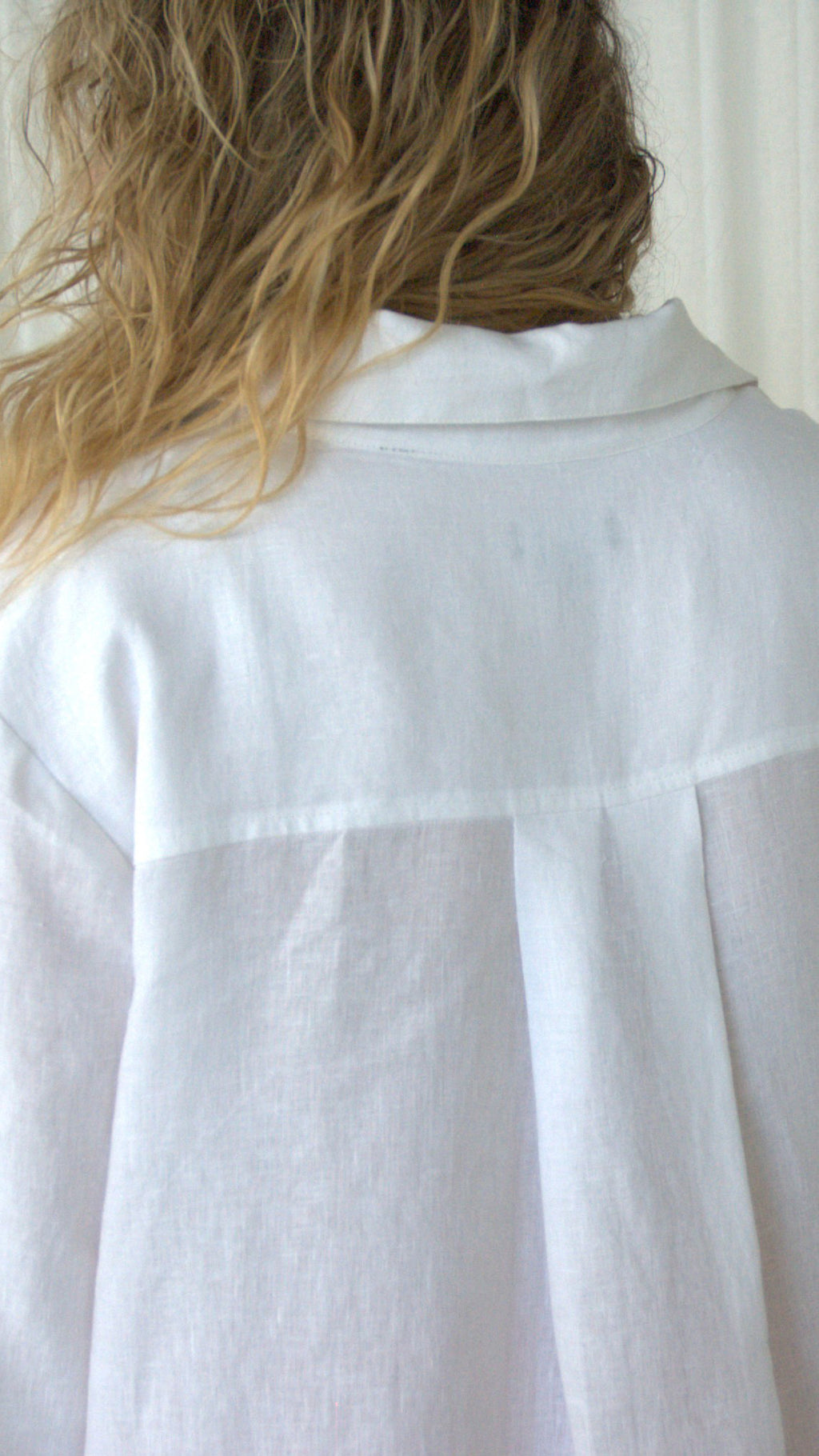 A close-up back view of a person wearing a white linen open collar & buttoned shirt, showing the fabric texture, back yoke & back pleat.