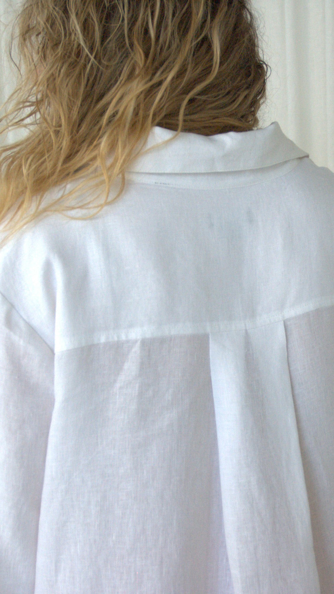 A close-up back view of a person wearing a white linen open collar & buttoned shirt, showing the fabric texture, back yoke & back pleat.