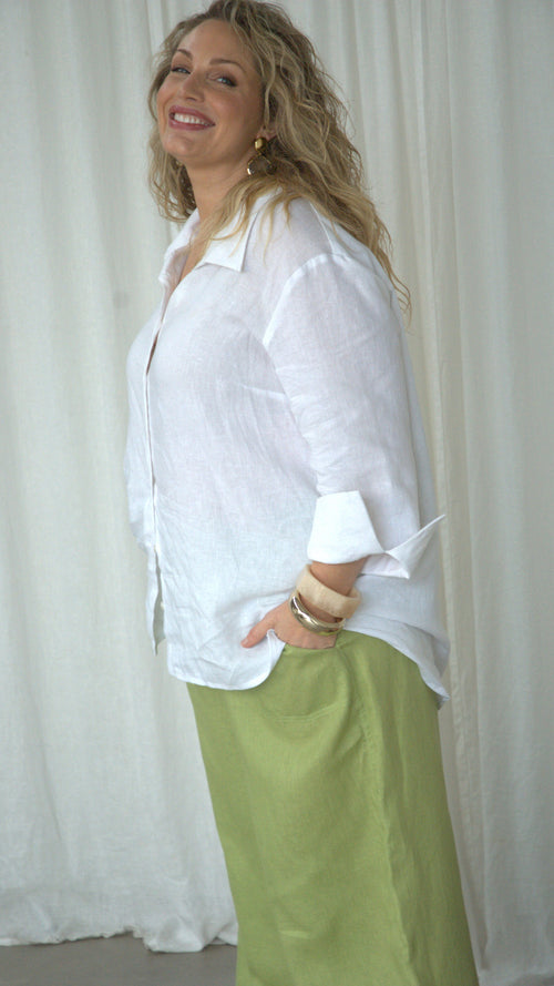 A woman wearing a white linen button shirt with sleeve cuffs and a light pear colour linen pants in front of a white curtain.