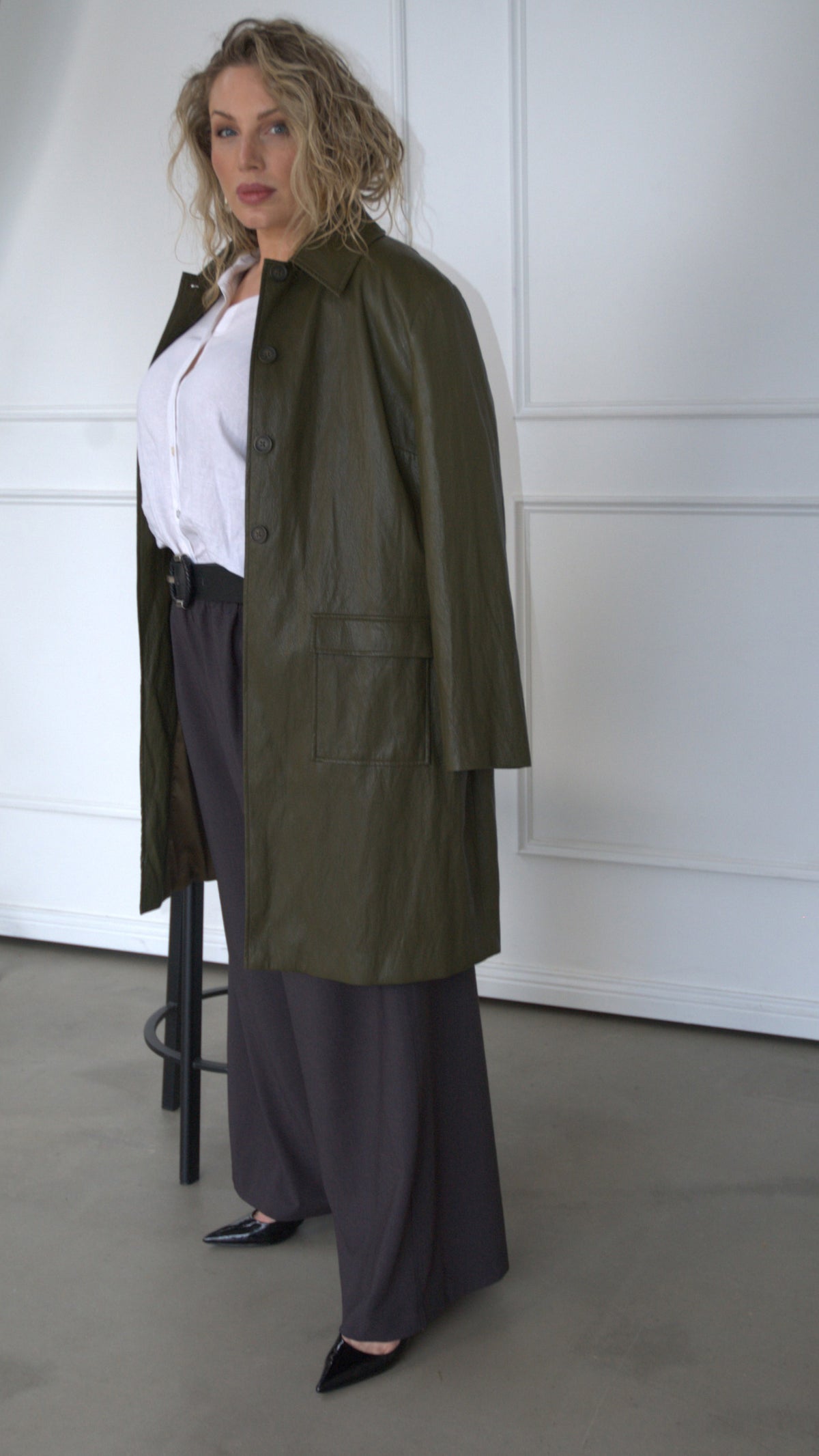 A woman models our knee length olive ARIA jacket over our LENNI white linen SHIRT and ABBI charcoal palazzo trousers, standing in a minimalist room with white paneled walls and concrete floor.