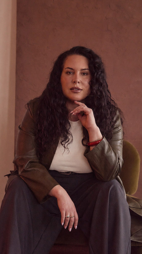 Model sitting on a chair wearing olive jacket, cream tee, charcoal pant, belt & red bangle