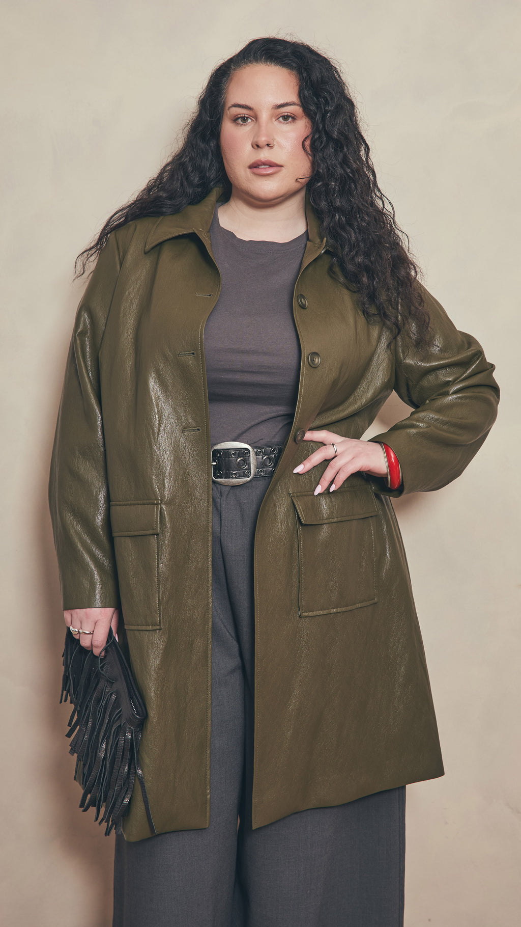 Modal wearing a olive leather effect jacket holding a black leather fringe  clutch on a beige background