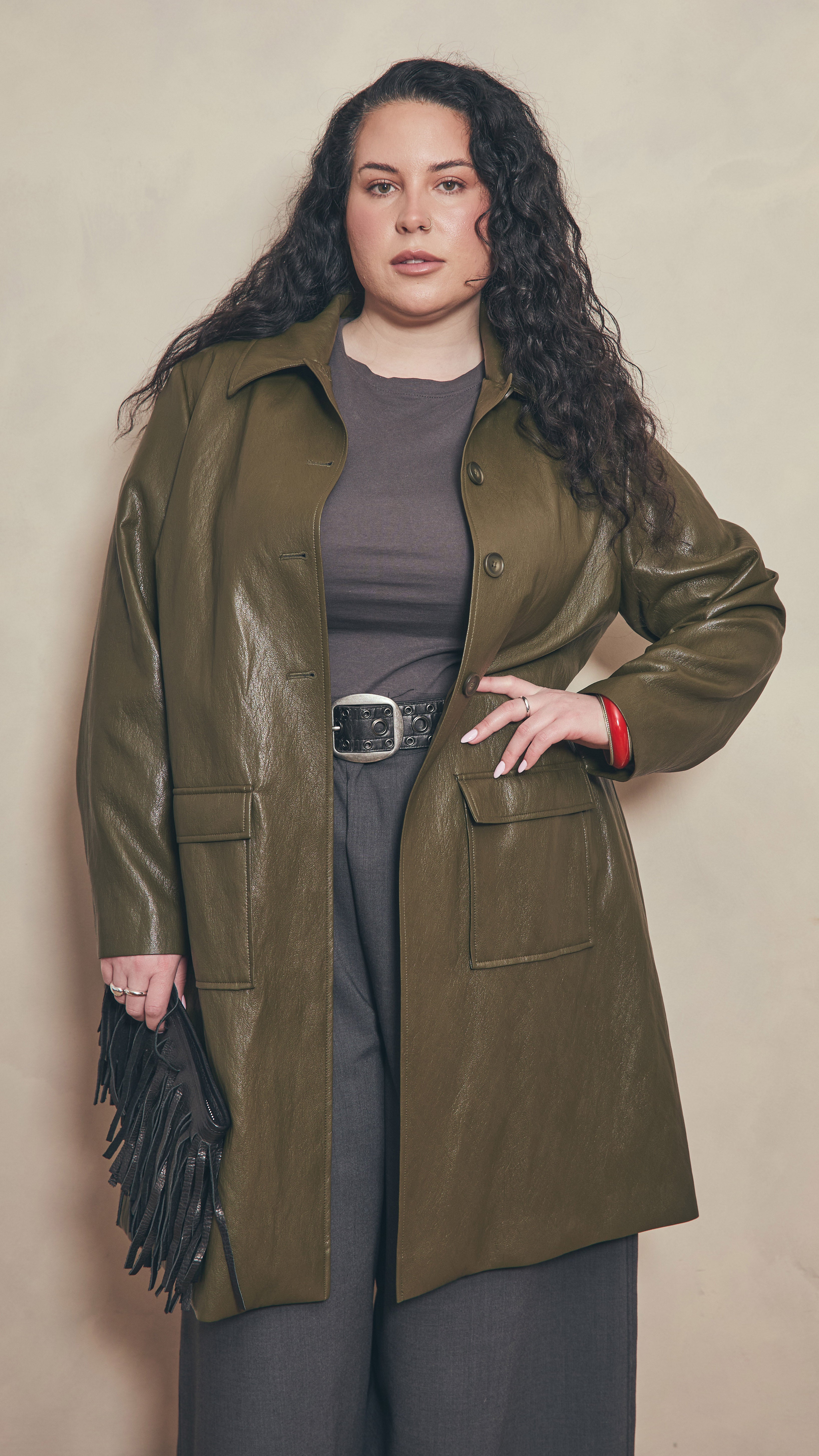 Modal wearing a olive leather effect jacket holding a black leather fringe  clutch on a beige background