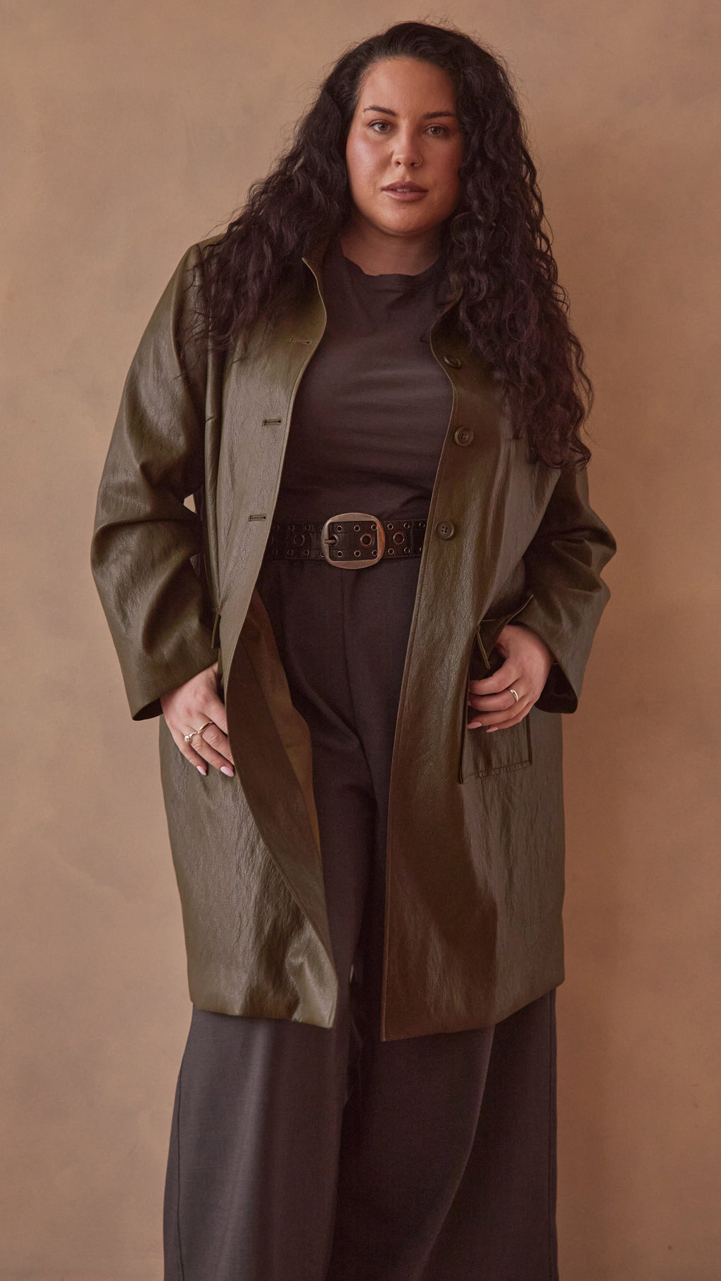 Modal wearing a olive green coat over a charcoal pants & top against a beige background