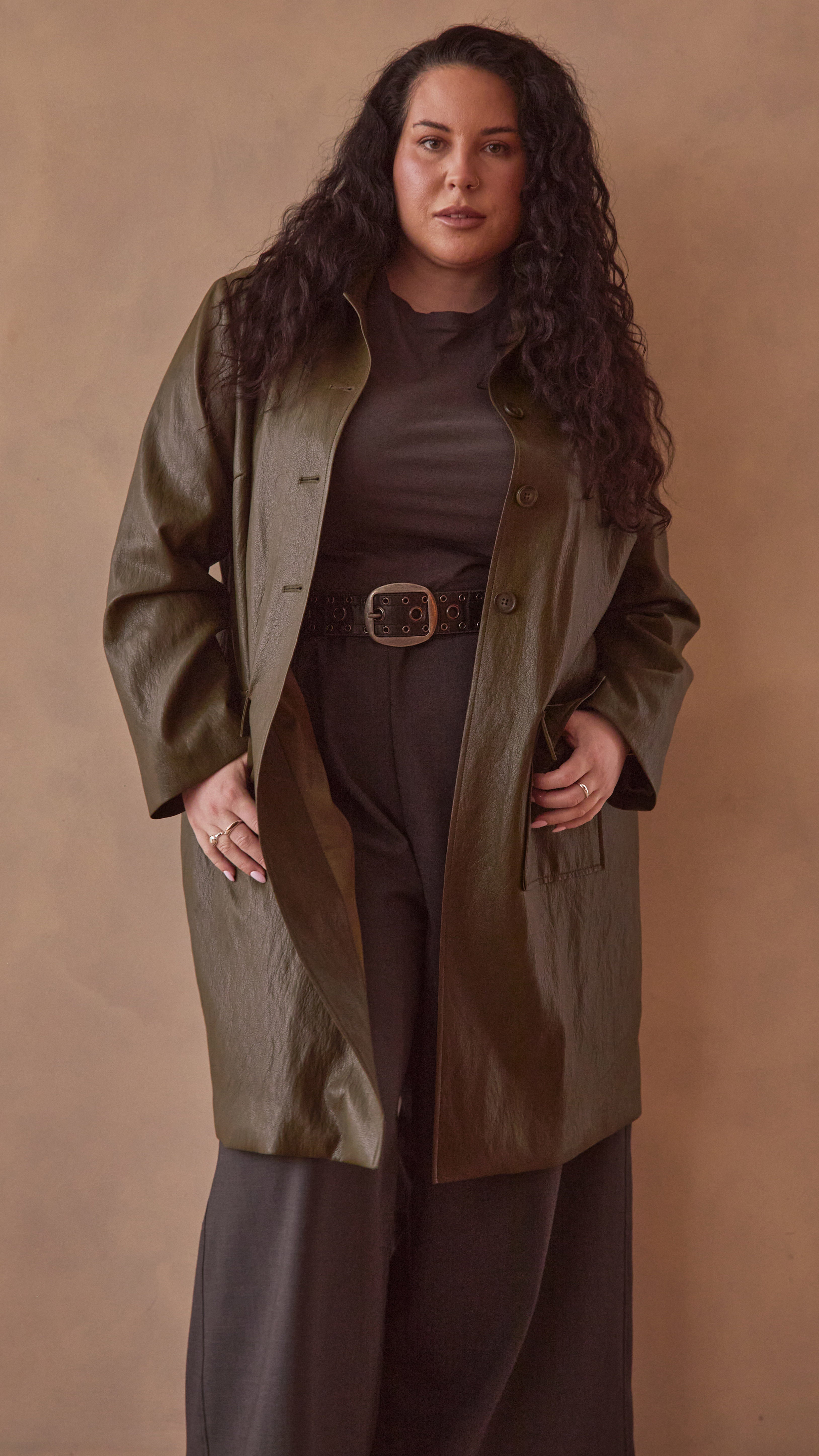 Modal wearing a olive green coat over a charcoal pants & top against a beige background