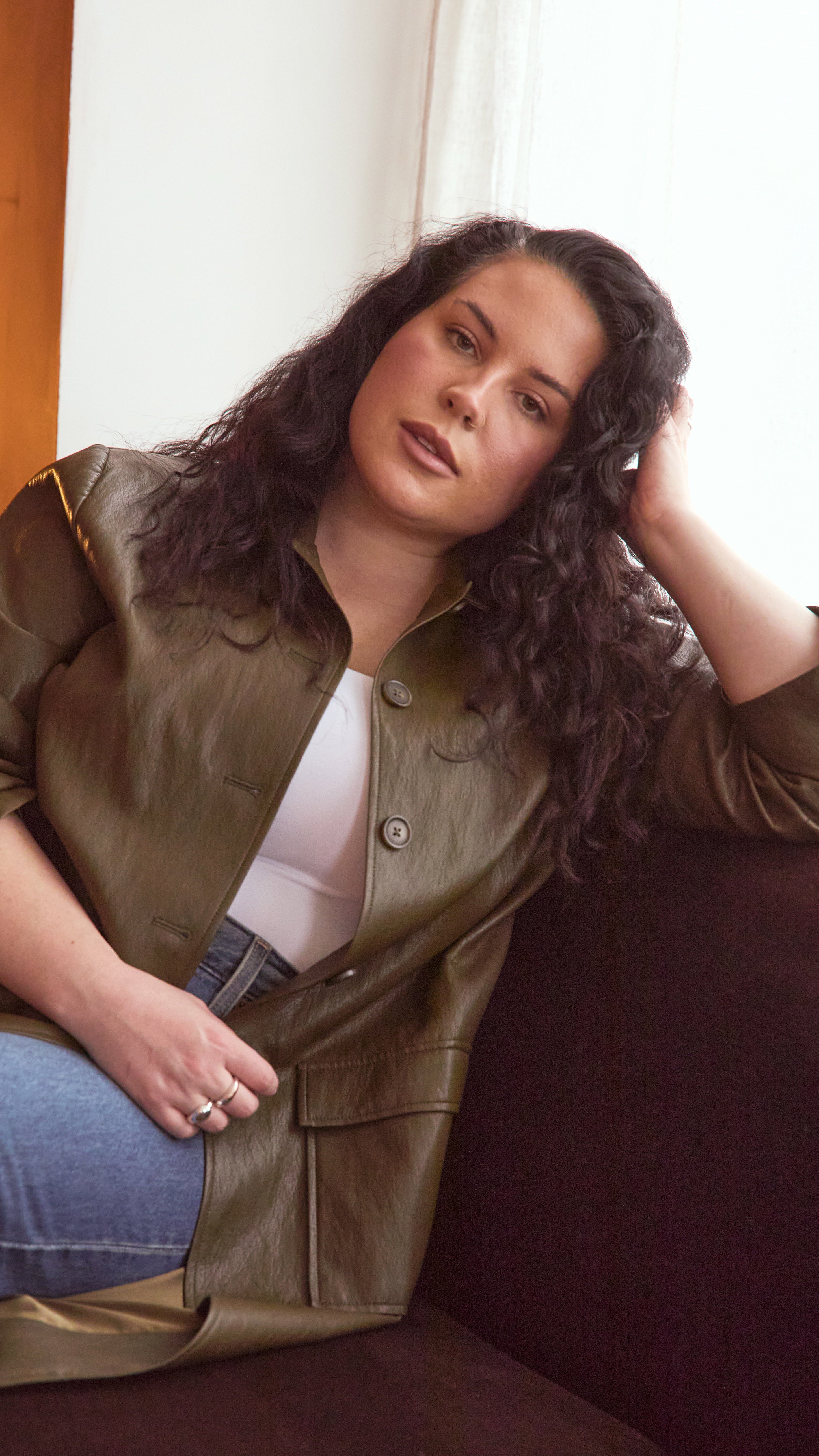 Modal wearing a olive jacket sitting inside on a burgundy couch.