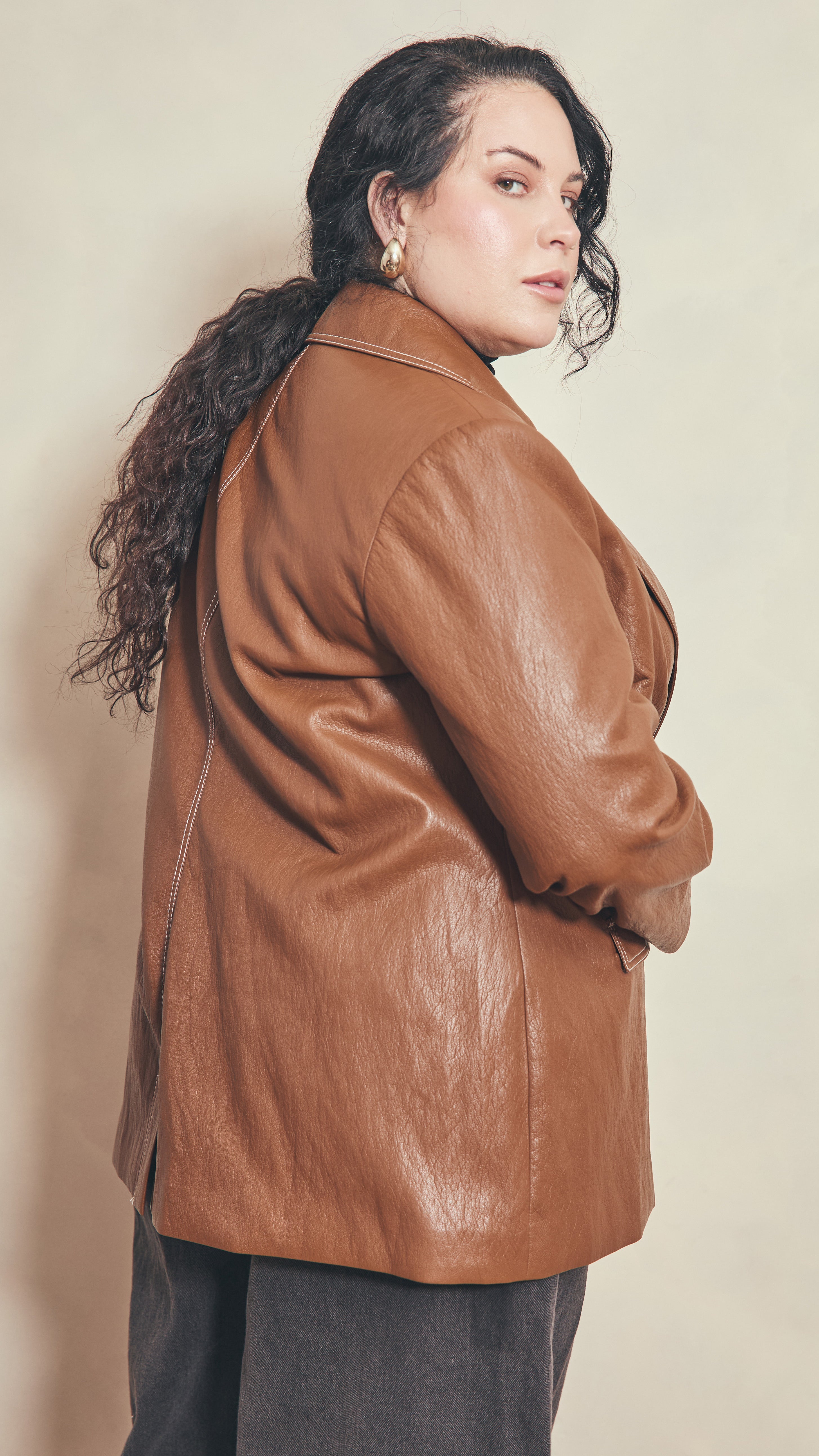 Side back view of modal wearing a burnt toffee colour leather effect jacket against a beige background