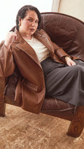 Modal sitting on a brown leather armchair wearing a burnt toffee colour jacket with a cream top and charcoal. coat.