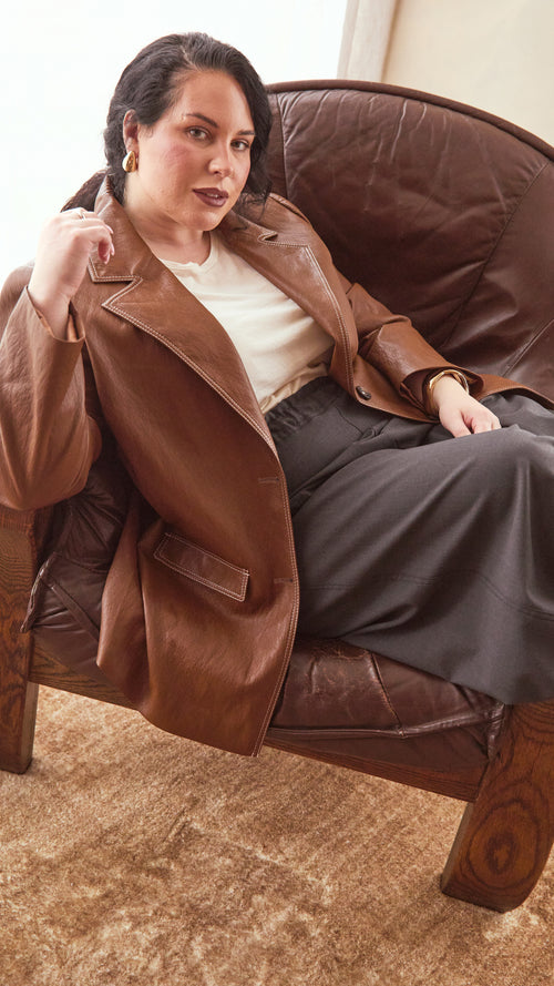 Modal sitting on a brown leather armchair wearing a burnt toffee colour jacket with a cream top and charcoal. coat.