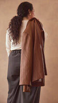 Model wearing charcoal pants with cream long sleeve top and burnt toffee jacket slung over her shoulder background