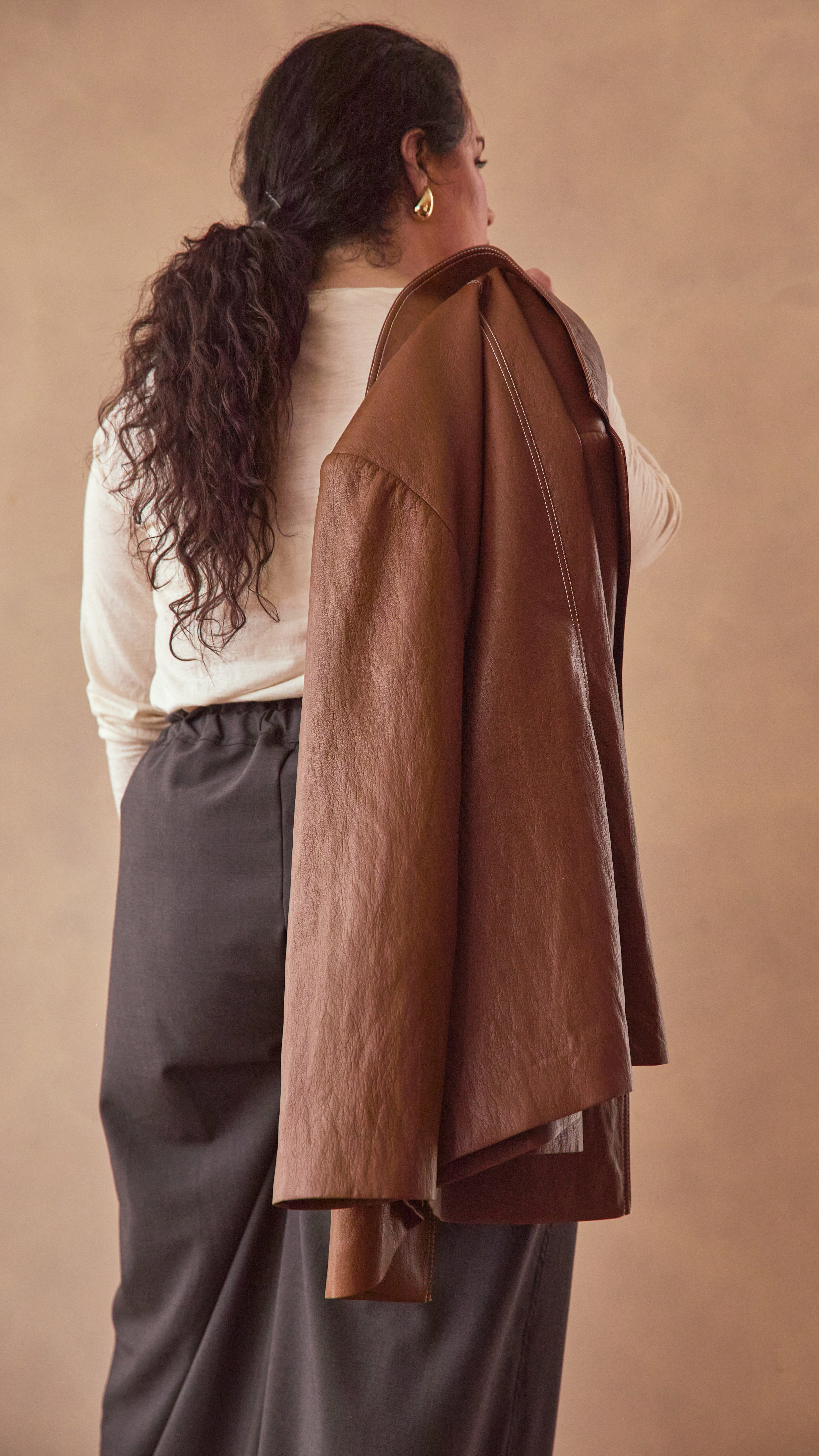 Model wearing charcoal pants with cream long sleeve top and burnt toffee jacket slung over her shoulder background