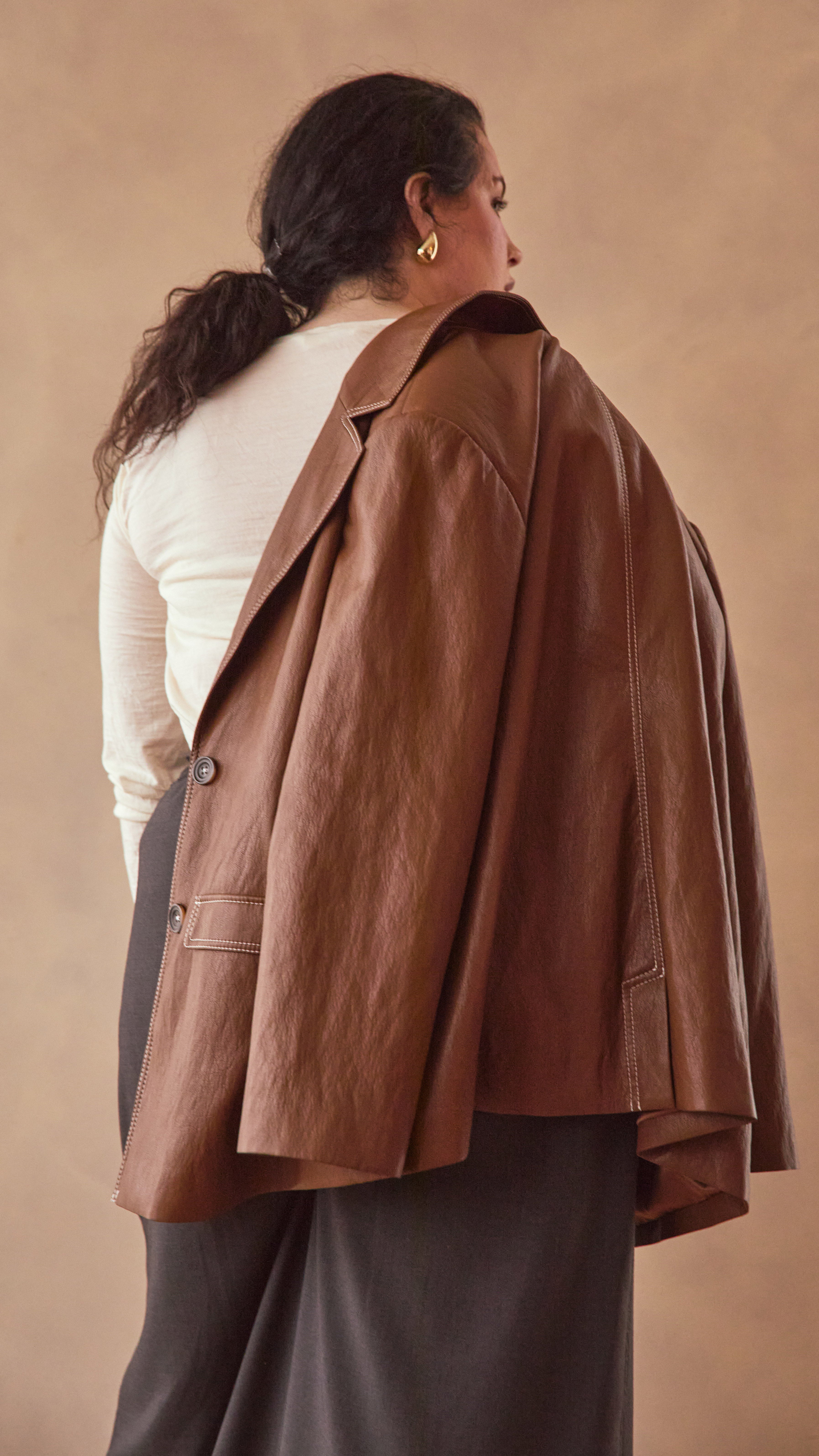 Back view of modal wearing a burnt toffee colour leather effect blazer against a beige background