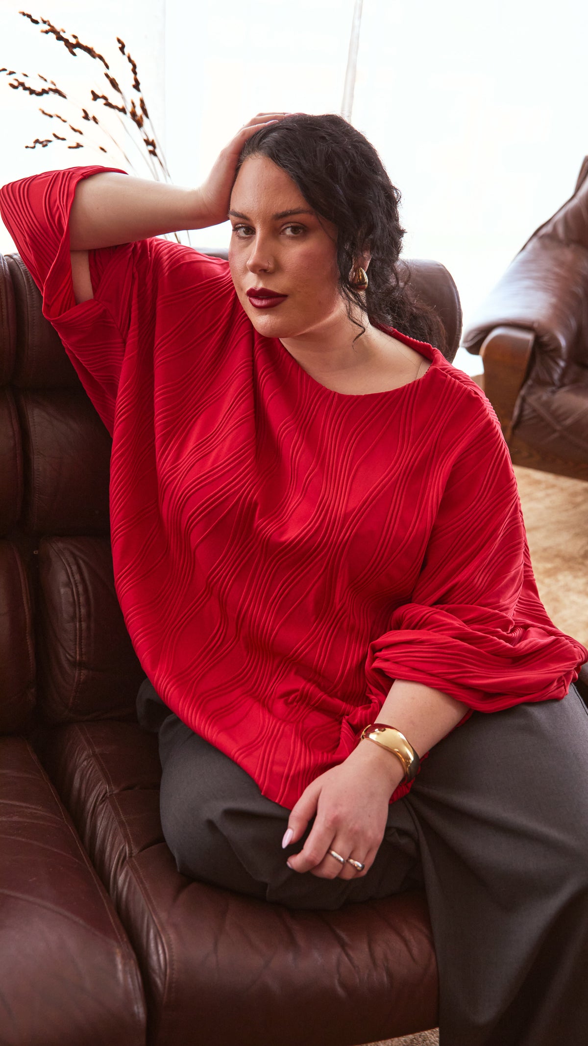 Editorial image. Modal in a red dolman top and charcoal pants, hand on head, sitting on a brown 80's leather couch. Sheer white curtains on the windows behind her & some dried flowers standing in a vase on the floor beind the couch.