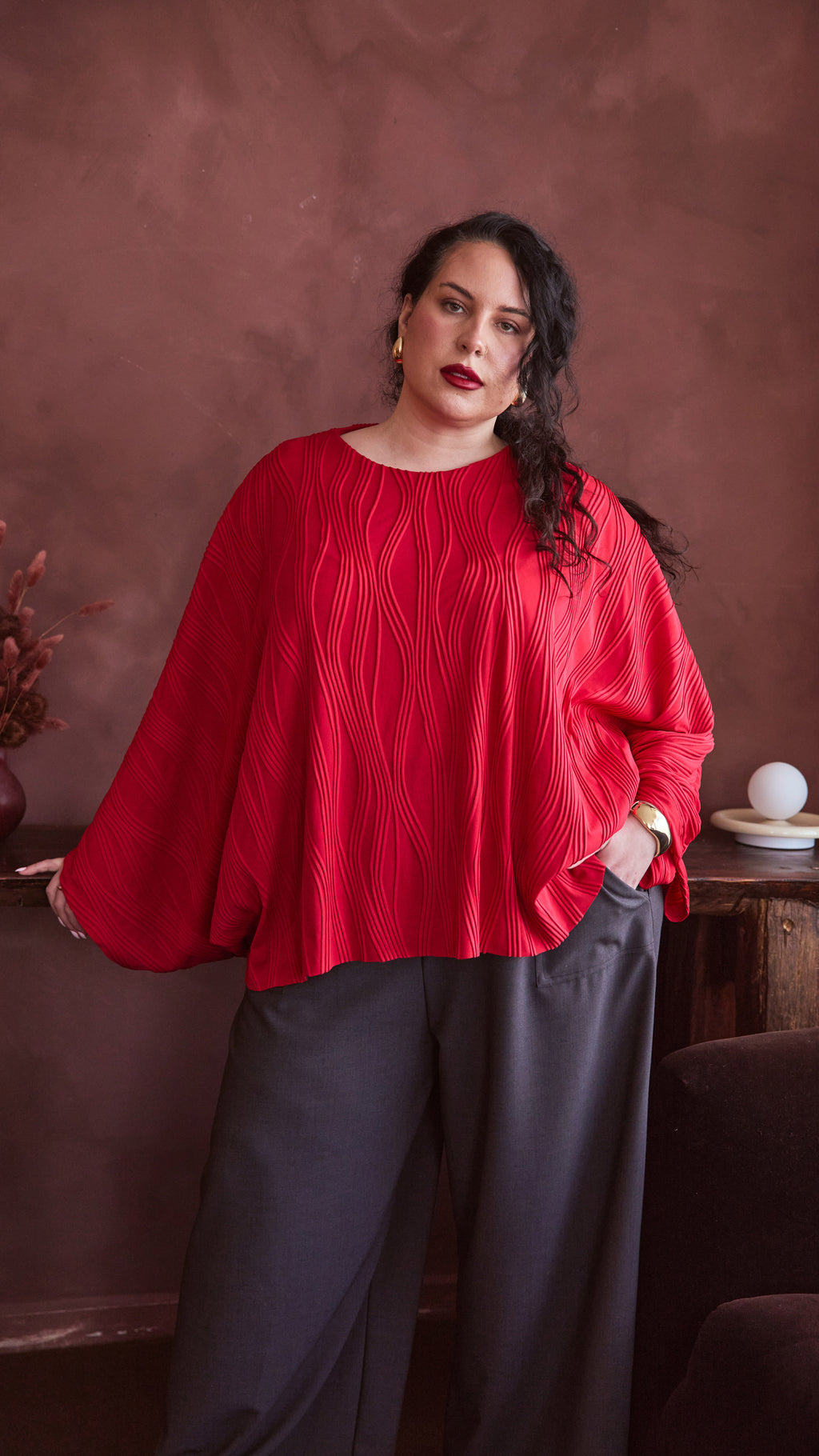 Modal standing against chunky wooden table, one hand in pocket the other holding the table. She is wearing a red dolman top and charcoal pants.