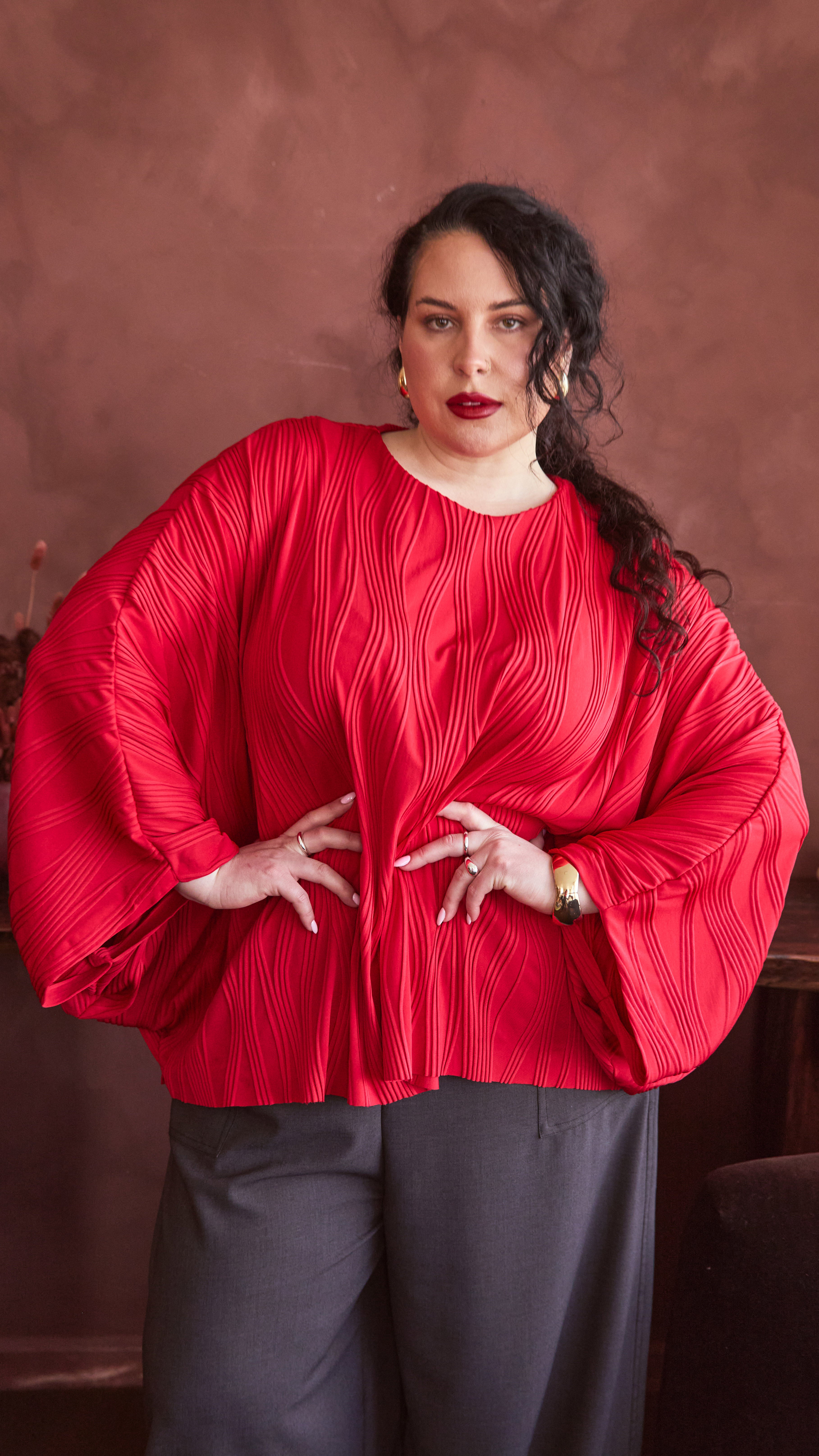 Modal wearing a red dolman top, with hands on hip. She has gold earrings, bangle and rings. The fabric on the top is a jacquard with irregular, vertical wave design. 
