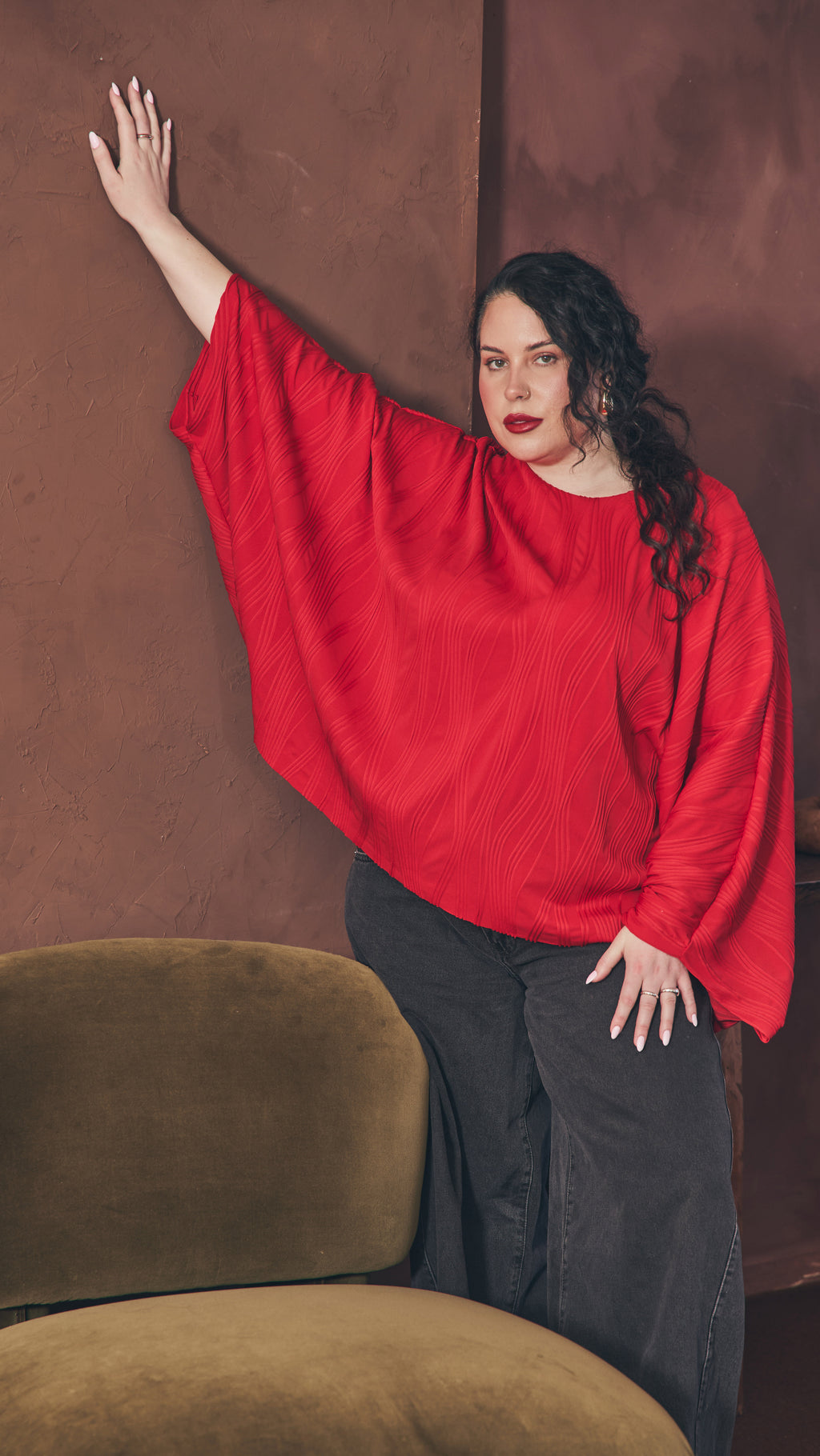 Modal standing next to olive velvet chair with one hand resting up against the  dark brown wall and the other resting on her thigh. Shows the oversize of the red dolman top.