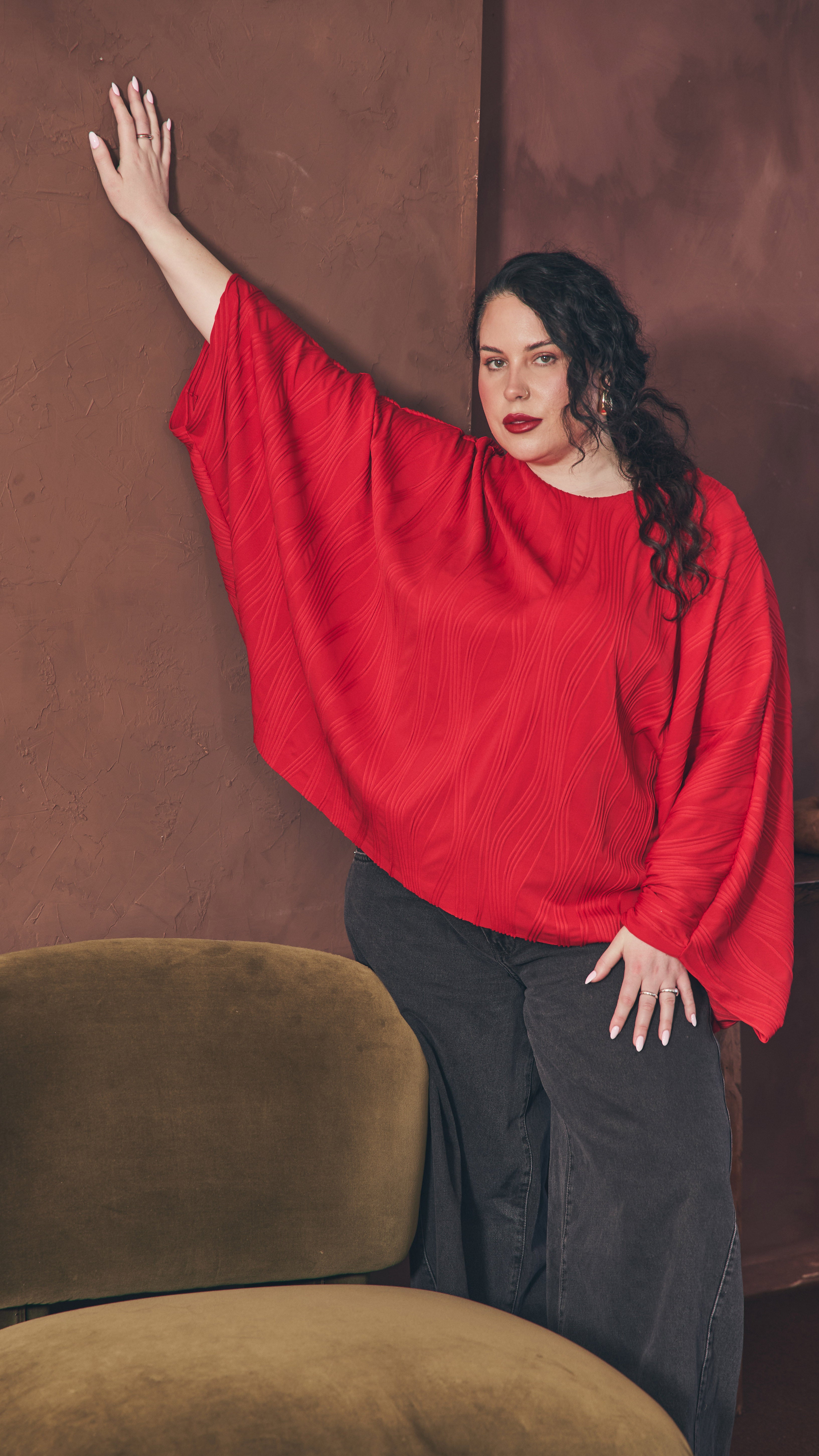 Modal standing next to olive velvet chair with one hand resting up against the  dark brown wall and the other resting on her thigh. Shows the oversize of the red dolman top.