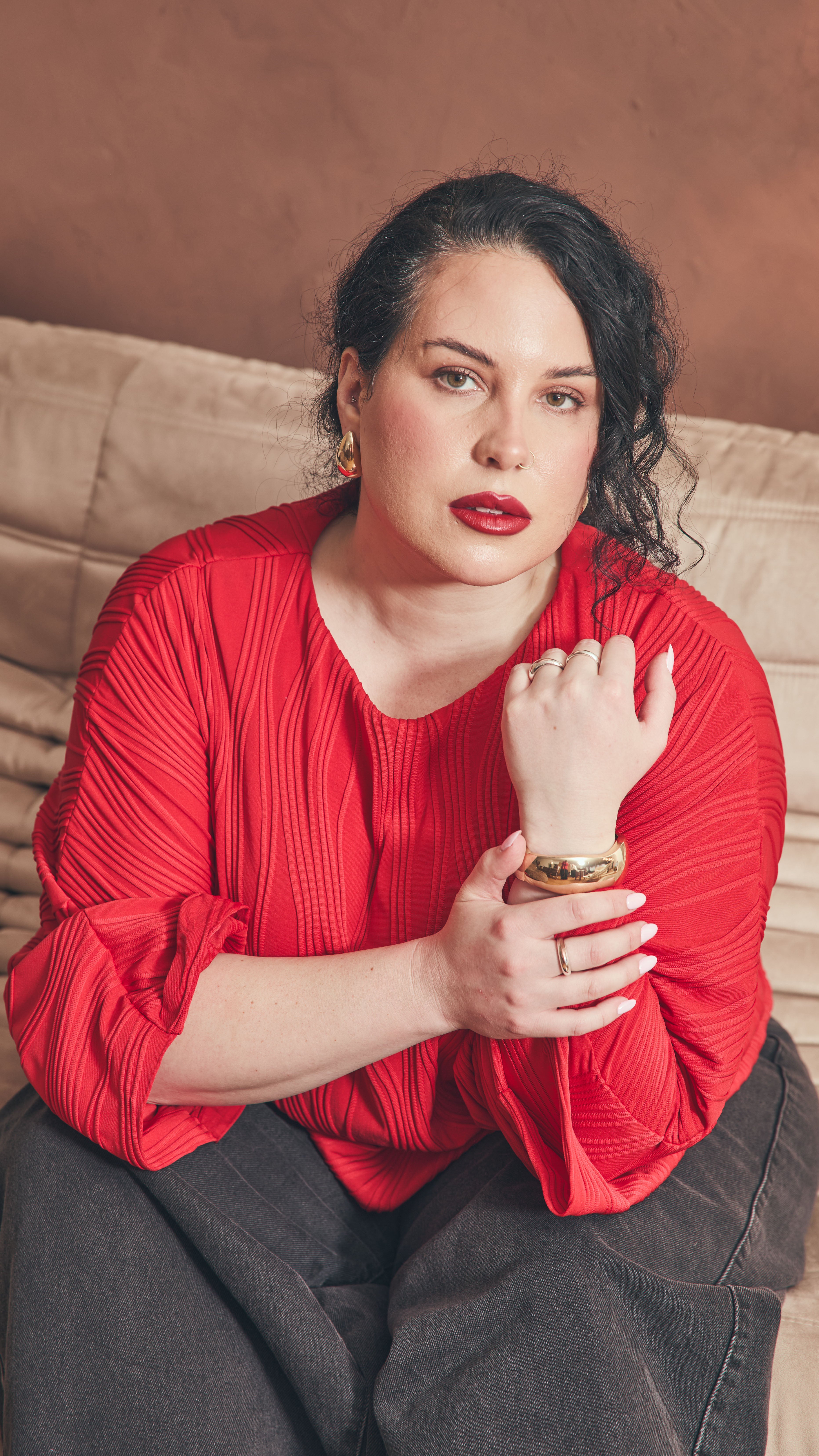 Modal sitting on beige leather couch wearing red dolman top, black denim barrel jeans, gold earrings and bangle holding her arm across her front. Wearing red lipstick that matches the top.